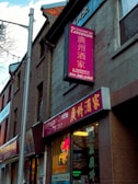 A street view of a building with a Cantonese restaurant. The signs, written in both English and Chinese, advertise the restaurant with bright colors. Neon lights are visible in the windows, and the architecture includes brickwork and a large pole.