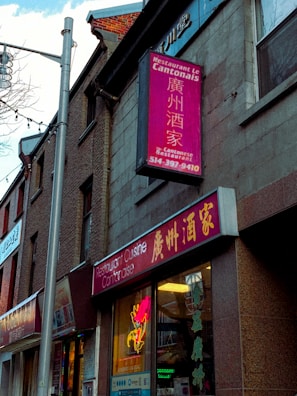 A street view of a building with a Cantonese restaurant. The signs, written in both English and Chinese, advertise the restaurant with bright colors. Neon lights are visible in the windows, and the architecture includes brickwork and a large pole.