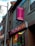 A street view of a building with a Cantonese restaurant. The signs, written in both English and Chinese, advertise the restaurant with bright colors. Neon lights are visible in the windows, and the architecture includes brickwork and a large pole.