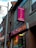 A street view of a building with a Cantonese restaurant. The signs, written in both English and Chinese, advertise the restaurant with bright colors. Neon lights are visible in the windows, and the architecture includes brickwork and a large pole.