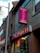 A street view of a building with a Cantonese restaurant. The signs, written in both English and Chinese, advertise the restaurant with bright colors. Neon lights are visible in the windows, and the architecture includes brickwork and a large pole.