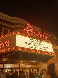 A theater marquee at night displays an event for Yves Tumor, Pretty Sick & Nation, with a date and time of May 12 at 7:00 PM. The neon sign reads 'Riviera' in bold red letters. A crowd of people is gathered below the marquee, suggesting an event taking place.