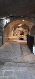 A dimly lit underground brick tunnel with archways leads to an area filled with ancient artifacts. Stone blocks, a large triangular object, and a framed picture are visible. The ambiance suggests it is part of an archaeological or historical site.