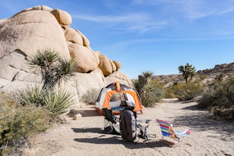 A desert campsite with an orange and white tent set up near large boulders and surrounded by desert vegetation, including Joshua trees. Two chairs and camping gear are placed in front of the tent under a clear blue sky.