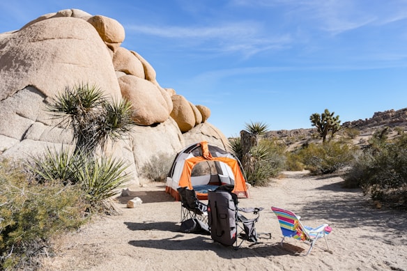 A desert campsite with an orange and white tent set up near large boulders and surrounded by desert vegetation, including Joshua trees. Two chairs and camping gear are placed in front of the tent under a clear blue sky.