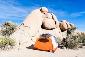 A desert landscape with large boulders, featuring an orange and white camping tent situated in the foreground. Surrounded by desert plants under a clear blue sky.