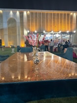A luxurious setting features a long, reflective table with a centerpiece of vibrant pink and red flowers in a metallic vase. The background shows a grand, illuminated building with a patterned facade and the words 'Delite Forever Orchid' above the entrance. Several people stand near the entrance, and the area is lit with warm, ambient lighting, creating an elegant atmosphere.