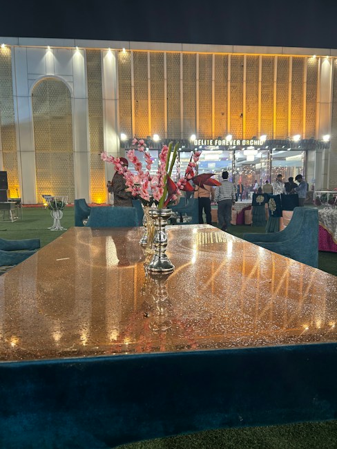 A luxurious setting features a long, reflective table with a centerpiece of vibrant pink and red flowers in a metallic vase. The background shows a grand, illuminated building with a patterned facade and the words 'Delite Forever Orchid' above the entrance. Several people stand near the entrance, and the area is lit with warm, ambient lighting, creating an elegant atmosphere.