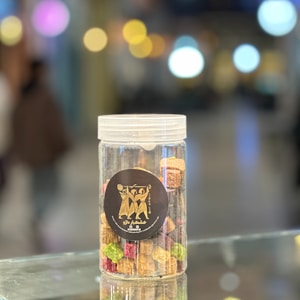 A transparent plastic jar filled with colorful rock candies, featuring a black circular label with artistic figures in gold. The background is blurred with bokeh lights, creating a festive and lively atmosphere.