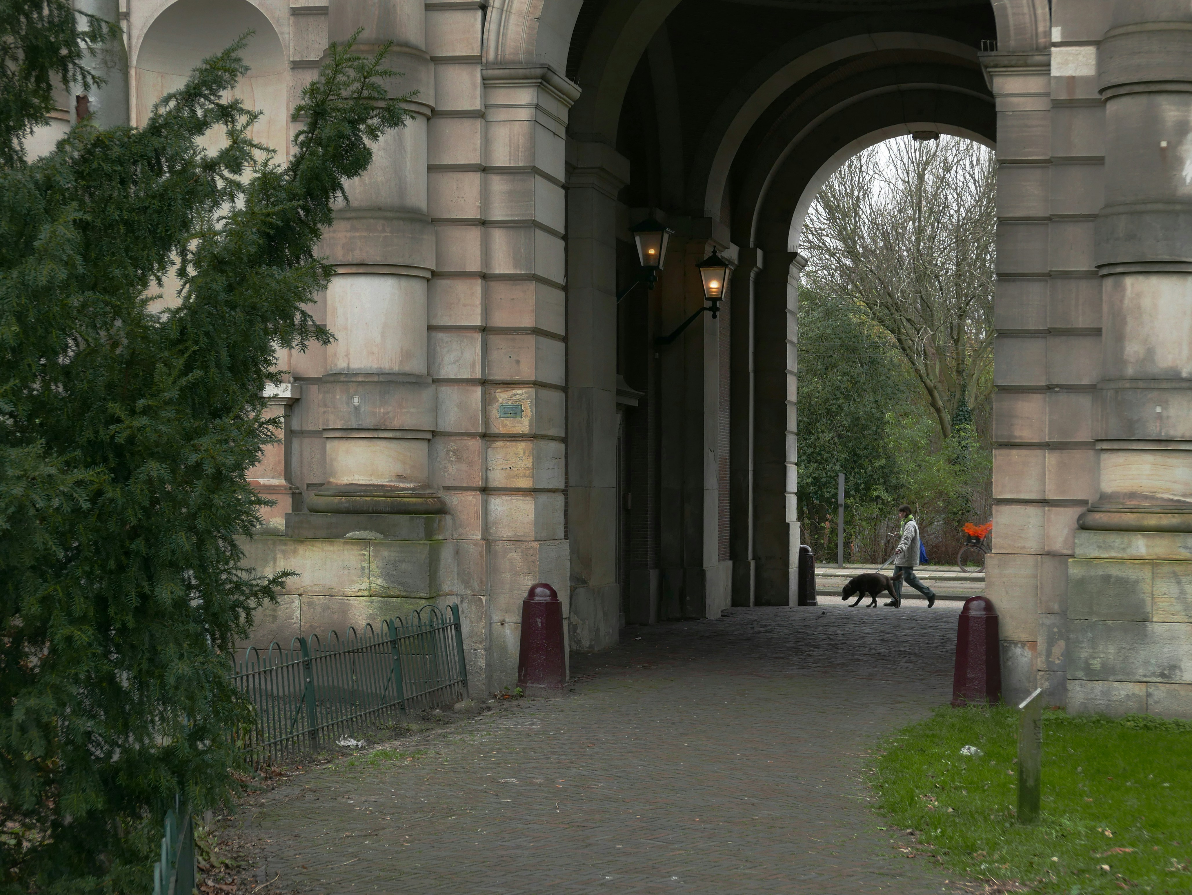 A look through image of the old Dutch city gate called Muiderpoort, in Amsterdam city, built in stone circa 1790. This arch replaced the old original city gate here. A man is just walking the dog in the look-through area. I like very much the stone textiure and pattern. Street photography of old cities in The Netherlands by Fons Heijnsbroek, 2023; Dutch photographer // Amsterdam in herfst, foto van de Muiderpoort, opgebouwd in natuursteen circa 1790. Een man laat net zijn hond uit. Fons Heijnsbroek: straat fotografie en oude architectuur in Nederland, in hoge resolutie, gratis foto.