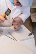 A person wearing a hat and glasses is engaged in stone carving, using a chisel and hammer on a marble slab. Several carving tools are visible around the workspace, which appears to be a well-lit studio.