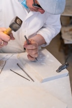 A person wearing a hat and glasses is engaged in stone carving, using a chisel and hammer on a marble slab. Several carving tools are visible around the workspace, which appears to be a well-lit studio.