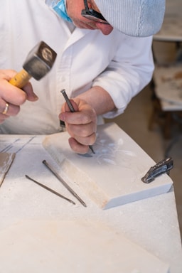 A person wearing a hat and glasses is engaged in stone carving, using a chisel and hammer on a marble slab. Several carving tools are visible around the workspace, which appears to be a well-lit studio.
