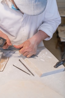 A person wearing a white shirt and a striped cap is carefully carving designs into a stone slab using a chisel and hammer. The workbench is covered in stone dust, and there are additional chisels lying nearby.