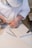 A person wearing a white shirt and a striped cap is carefully carving designs into a stone slab using a chisel and hammer. The workbench is covered in stone dust, and there are additional chisels lying nearby.