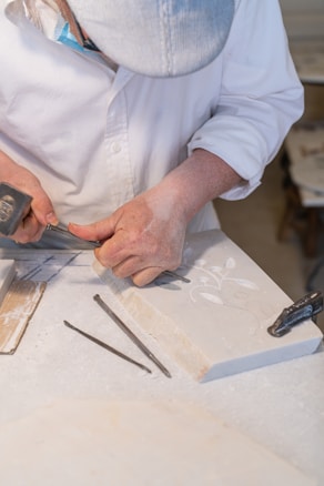 A person wearing a white shirt and a striped cap is carefully carving designs into a stone slab using a chisel and hammer. The workbench is covered in stone dust, and there are additional chisels lying nearby.