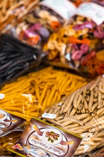 Various types of packaged pasta are displayed, featuring a mix of colors and shapes. The packages have clear labels, and the pasta includes traditional as well as colorful varieties made with different ingredients.