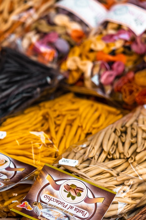 Various types of packaged pasta are displayed, featuring a mix of colors and shapes. The packages have clear labels, and the pasta includes traditional as well as colorful varieties made with different ingredients.