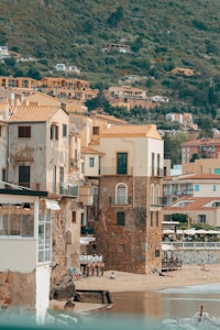 A picturesque seaside village with rustic stone buildings and yellow-orange rooftops nestled against a lush green hillside. The scene captures a few people relaxing on a small sandy beach by the calm sea. The village comprises several homes and small structures, with a mix of residential and commercial establishments visible.
