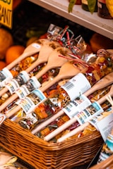 A basket contains bags of colorful pasta displayed with wooden spoons. The pasta is packaged in clear bags adorned with labels that have images and text. The display appears to be part of a market stall or shop, with other produce like oranges visible in the background.