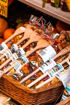 A basket contains bags of colorful pasta displayed with wooden spoons. The pasta is packaged in clear bags adorned with labels that have images and text. The display appears to be part of a market stall or shop, with other produce like oranges visible in the background.