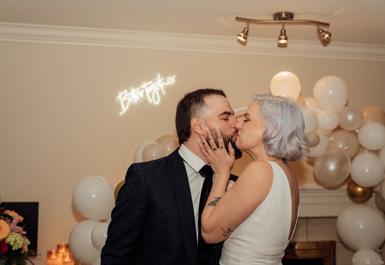 A couple shares a romantic kiss surrounded by white and gold balloons. A neon sign reading 'Better Together' is illuminated on the wall behind them. The setting appears festive, likely at a celebration or wedding.