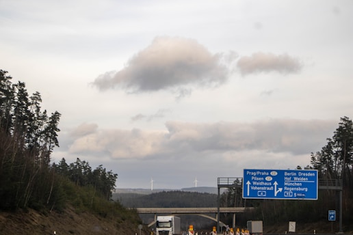 A highway scene with a directional road sign pointing towards multiple European cities. Tall trees line both sides of the road, and there are wind turbines visible in the distance. The sky is cloudy with a mix of grey and white clouds.
