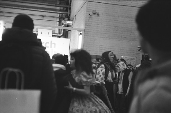 A black and white photo depicting a group of people in a crowded indoor setting, with some individuals in traditional or cultural attire. The background includes a brick wall and signage. The scene appears dynamic, with people interacting.