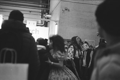 A black and white photo depicting a group of people in a crowded indoor setting, with some individuals in traditional or cultural attire. The background includes a brick wall and signage. The scene appears dynamic, with people interacting.