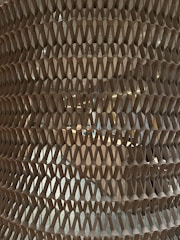 A close-up view of an intricate, honeycomb-like cardboard or paper structure with repeating triangular patterns. The structure appears semi-transparent, allowing a glimpse of what seems to be an interior space or objects behind it with some light filtering through.