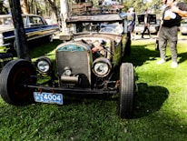 A vintage car with a rusty exterior and exposed engine parts is parked on a grassy area. It features large round headlights and a distinctive front grille, with a blue license plate on the front. The car has a skull ornament and other custom decorations, giving it a unique, personalized appearance. In the background, there are other classic cars and several people standing around, some inspecting the vehicles.
