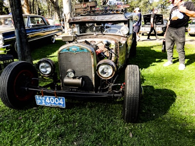 A vintage car with a rusty exterior and exposed engine parts is parked on a grassy area. It features large round headlights and a distinctive front grille, with a blue license plate on the front. The car has a skull ornament and other custom decorations, giving it a unique, personalized appearance. In the background, there are other classic cars and several people standing around, some inspecting the vehicles.