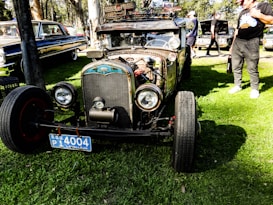 A vintage car with a rusty exterior and exposed engine parts is parked on a grassy area. It features large round headlights and a distinctive front grille, with a blue license plate on the front. The car has a skull ornament and other custom decorations, giving it a unique, personalized appearance. In the background, there are other classic cars and several people standing around, some inspecting the vehicles.