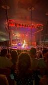 A circus performance takes place under a large tent. The stage is brightly illuminated with orange and red lights. A performer appears to be handling fire in front of an engaged audience. The background features an elaborate lighting setup with various colors.