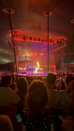 A circus performance takes place under a large tent. The stage is brightly illuminated with orange and red lights. A performer appears to be handling fire in front of an engaged audience. The background features an elaborate lighting setup with various colors.