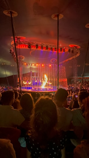 A circus performance takes place under a large tent. The stage is brightly illuminated with orange and red lights. A performer appears to be handling fire in front of an engaged audience. The background features an elaborate lighting setup with various colors.