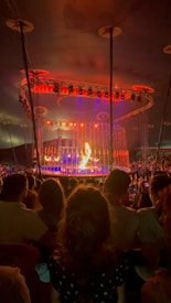 A circus performance takes place under a large tent. The stage is brightly illuminated with orange and red lights. A performer appears to be handling fire in front of an engaged audience. The background features an elaborate lighting setup with various colors.