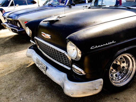 A classic black Chevrolet car is parked next to another vintage vehicle. The Chevrolet has a shiny chrome grille and distinctive round headlights. The metal parts of the wheels are polished, and the license plate is visible at the front. The background shows parts of another vintage car with a white and blue paint job.
