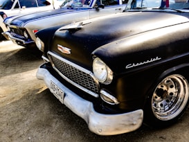 A classic black Chevrolet car is parked next to another vintage vehicle. The Chevrolet has a shiny chrome grille and distinctive round headlights. The metal parts of the wheels are polished, and the license plate is visible at the front. The background shows parts of another vintage car with a white and blue paint job.