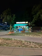 A flower shop is visible, brightly illuminated with green lights against a nighttime backdrop. The sign reads 'KWIACIARNIA 24h', indicating it's open 24 hours. The shop is adorned with various plants and greenery, and is surrounded by trees and grass.