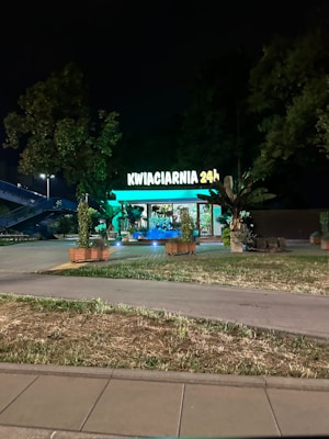 A flower shop is visible, brightly illuminated with green lights against a nighttime backdrop. The sign reads 'KWIACIARNIA 24h', indicating it's open 24 hours. The shop is adorned with various plants and greenery, and is surrounded by trees and grass.