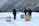 A group of people are walking on a frozen landscape, pulling sleds loaded with supplies. The environment is cold and harsh, with rugged rocky terrain surrounding the icy path. The people are dressed in heavy winter clothing, indicating severe weather conditions.