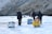 A group of people are walking on a frozen landscape, pulling sleds loaded with supplies. The environment is cold and harsh, with rugged rocky terrain surrounding the icy path. The people are dressed in heavy winter clothing, indicating severe weather conditions.