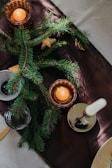 Sprigs of pine lay elegantly on a dark table runner, complemented by amber-hued candle holders casting a warm glow. A white unlit candle stands in a brass holder, adding a touch of elegance.