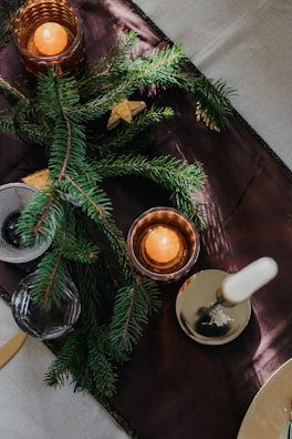 Sprigs of pine lay elegantly on a dark table runner, complemented by amber-hued candle holders casting a warm glow. A white unlit candle stands in a brass holder, adding a touch of elegance.