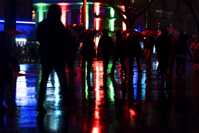 Silhouetted figures moving through a rain-soaked cityscape under glowing street lamps.
