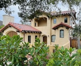 A two-story Mediterranean-style house with a terracotta roof and beige stucco walls. The building features black-framed windows and a wooden front door. It's surrounded by lush greenery, including trees and shrubs.
