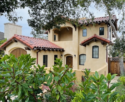 A two-story Mediterranean-style house with a terracotta roof and beige stucco walls. The building features black-framed windows and a wooden front door. It's surrounded by lush greenery, including trees and shrubs.