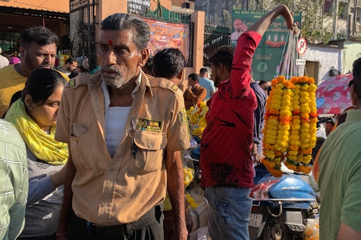 A busy street scene with a security guard wearing a uniform and a red mark on his forehead, surrounded by people. A vendor in a red shirt holds up several garlands made of yellow and orange flowers. A motorcycle covered in marigold garlands is nearby. The background shows a crowded market area with various stalls and street signs.