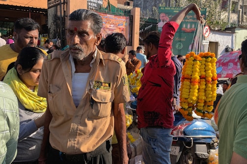 A busy street scene with a security guard wearing a uniform and a red mark on his forehead, surrounded by people. A vendor in a red shirt holds up several garlands made of yellow and orange flowers. A motorcycle covered in marigold garlands is nearby. The background shows a crowded market area with various stalls and street signs.